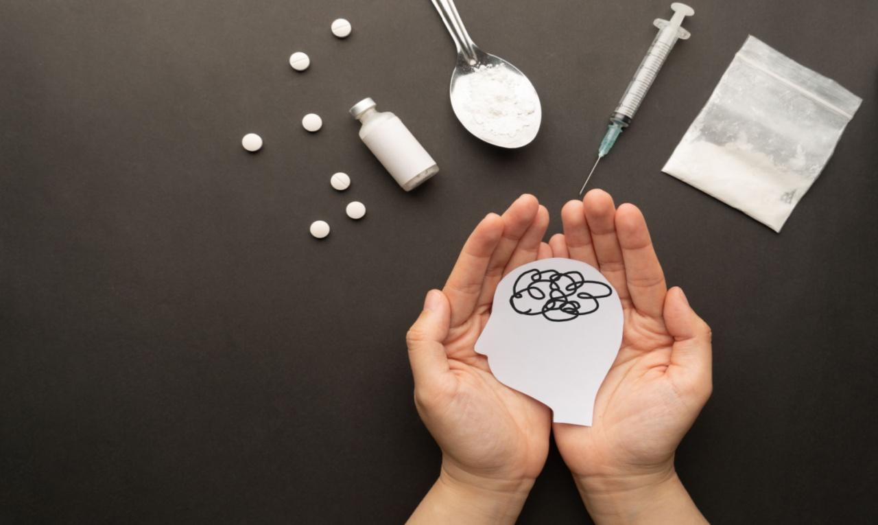 M&atilde;os segurando um recorte de papel em formato de c&eacute;rebro, com subst&acirc;ncias e seringa ao fundo