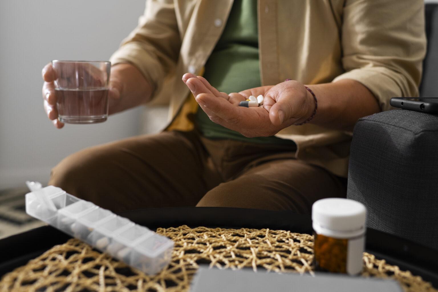 Homem sentado segurando comprimidos e um copo de água, com organizador semanal de medicamentos ao lado