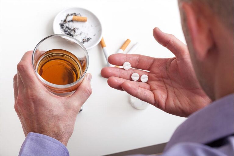 Mão segurando comprimidos ao lado de um copo de bebida alcoólica e cigarros