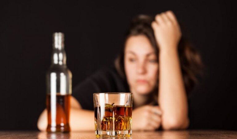 Mulher com a cabe&ccedil;a apoiada na m&atilde;o diante de um copo e uma garrafa de bebida alco&oacute;lica