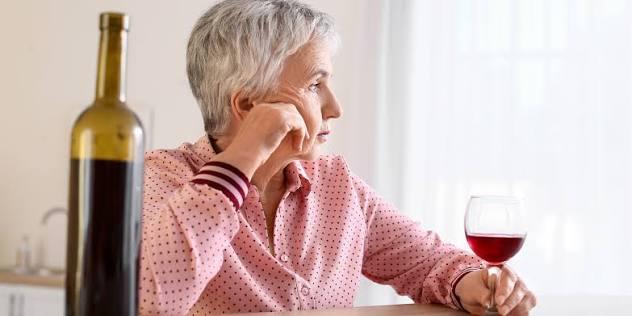 Mulher idosa sentada &agrave; mesa ao lado de uma garrafa e de uma ta&ccedil;a de vinho, olhando para a janela